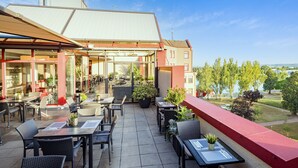 Een terras buiten met tafels en stoelen, een overdekte zithoek, en uitzicht op een meer en bomen.