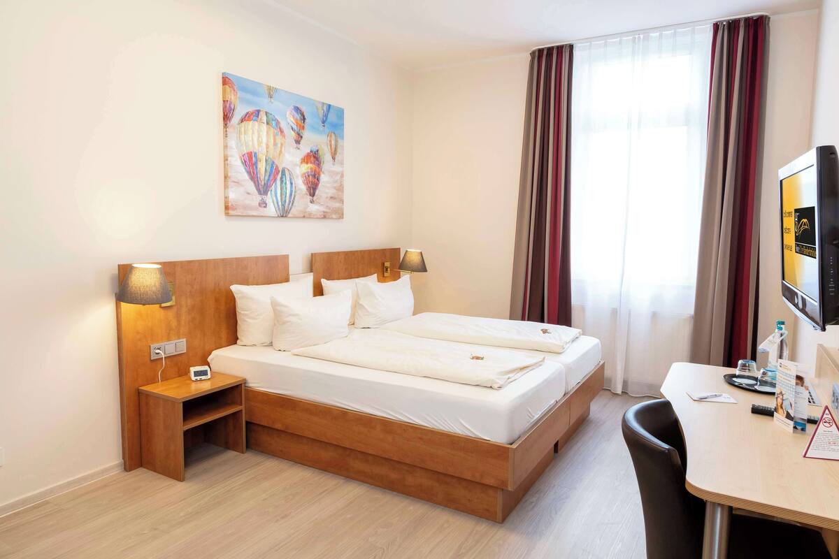Una habitación de hotel con una cama de madera, dos mesitas de noche, un escritorio con una silla, un televisor y un cuadro de globos aerostáticos.