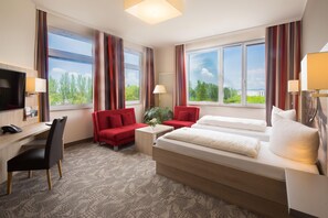 Una habitación de hotel con una cama, dos sillas rojas, un escritorio con teléfono y ventanas grandes con cortinas.