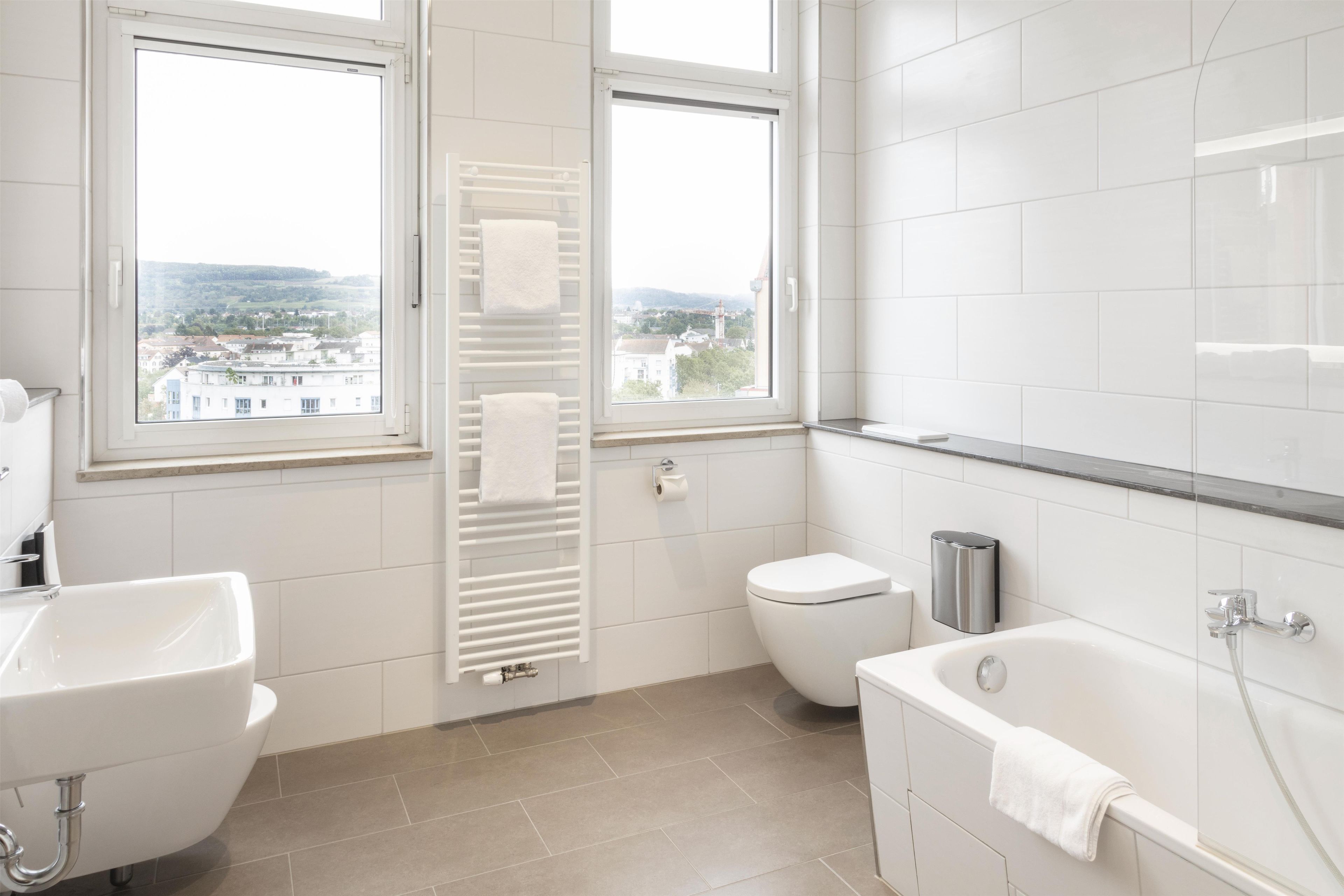 A modern bathroom with a bathtub, toilet, and sink. There are two windows providing a view of the cityscape.