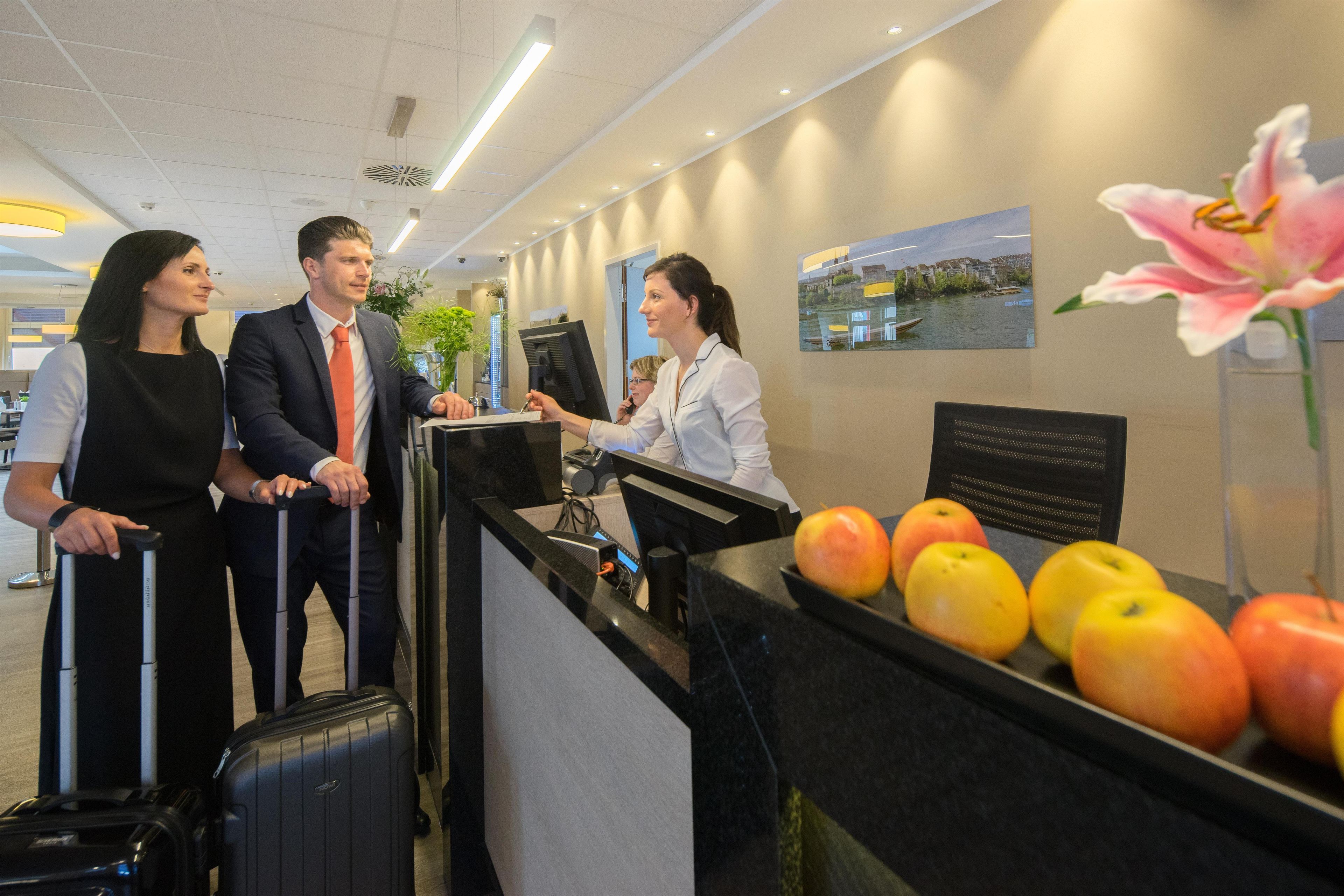 A reception area with a counter, a desk, and a wall-mounted display. There are apples on the counter and a flower vase.