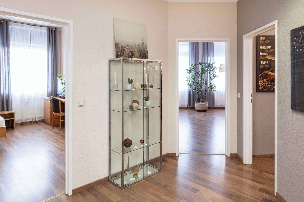 Una habitación amplia con suelo de madera, un aparador de vidrio, un área de comedor con mesa y sillas, y una planta grande en maceta.