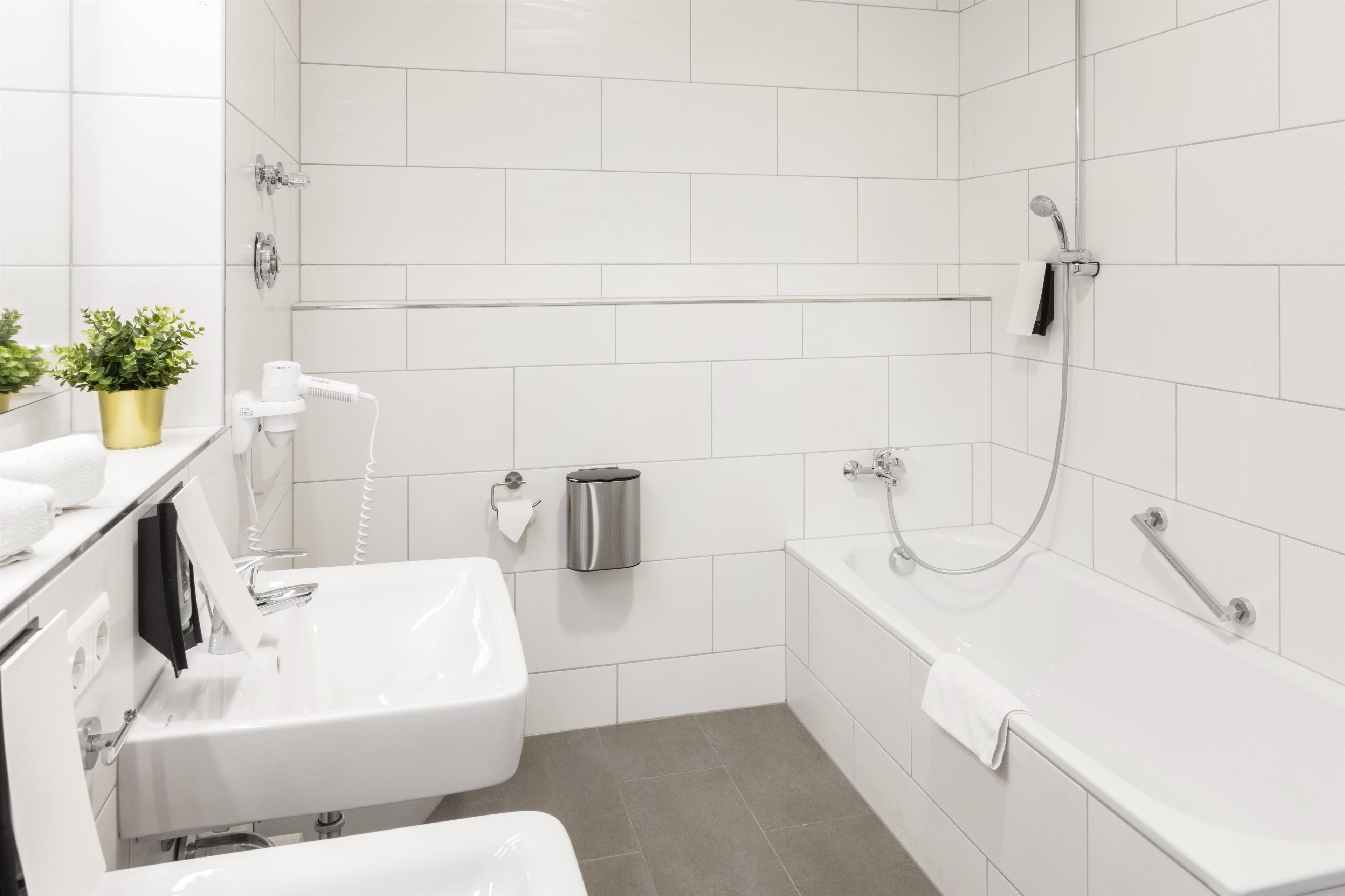 Un baño moderno con un lavabo, inodoro y bañera blancos. Hay un perchero, un cubo de basura y una pequeña planta en maceta sobre el mostrador.