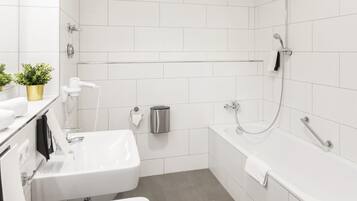 Un baño moderno con un lavabo, inodoro y bañera blancos. Hay un perchero, un cubo de basura y una pequeña planta en maceta sobre el mostrador.