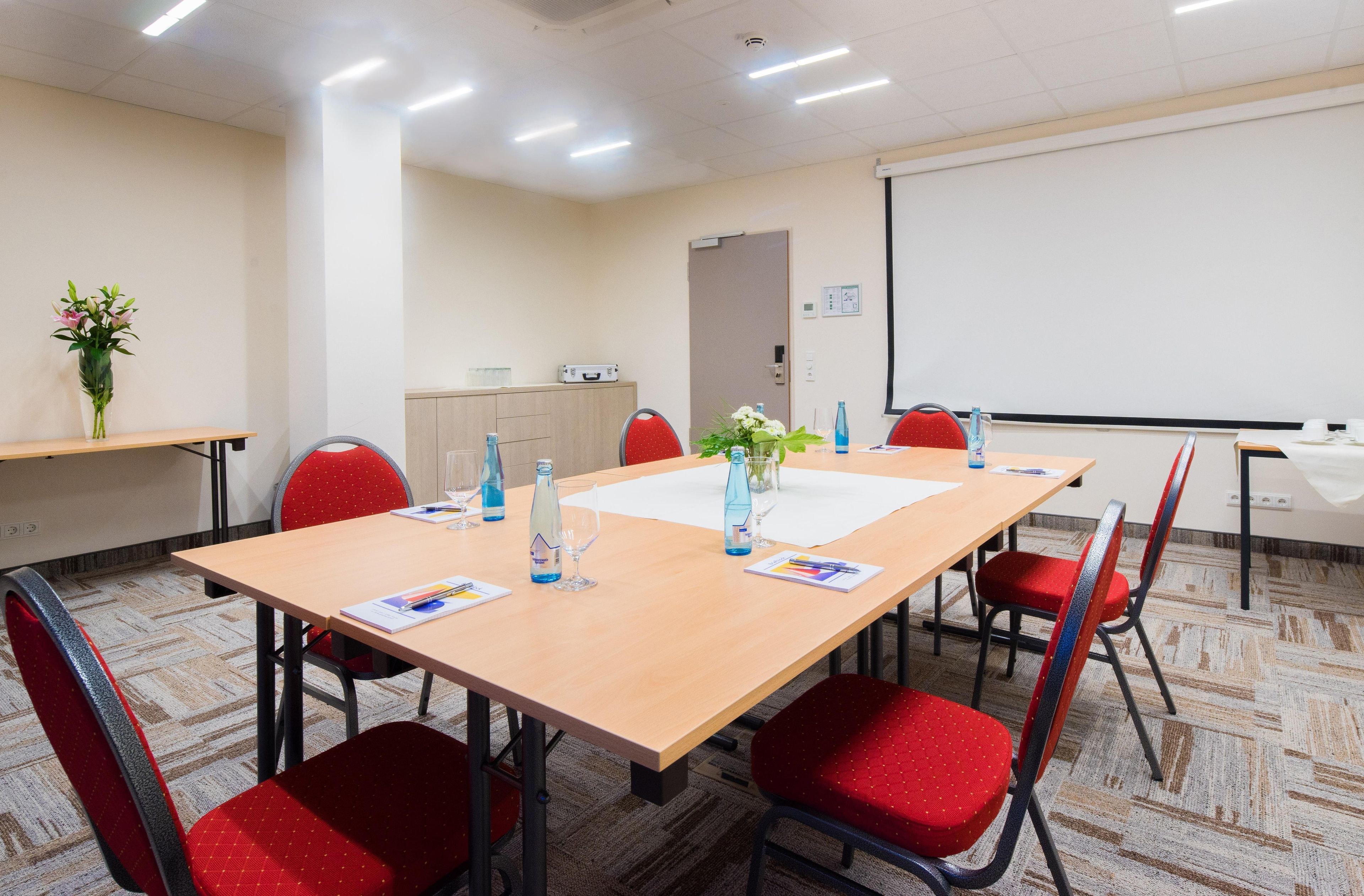 Une salle de conférence avec une longue table, des chaises rouges, un écran de projection et un tableau blanc.