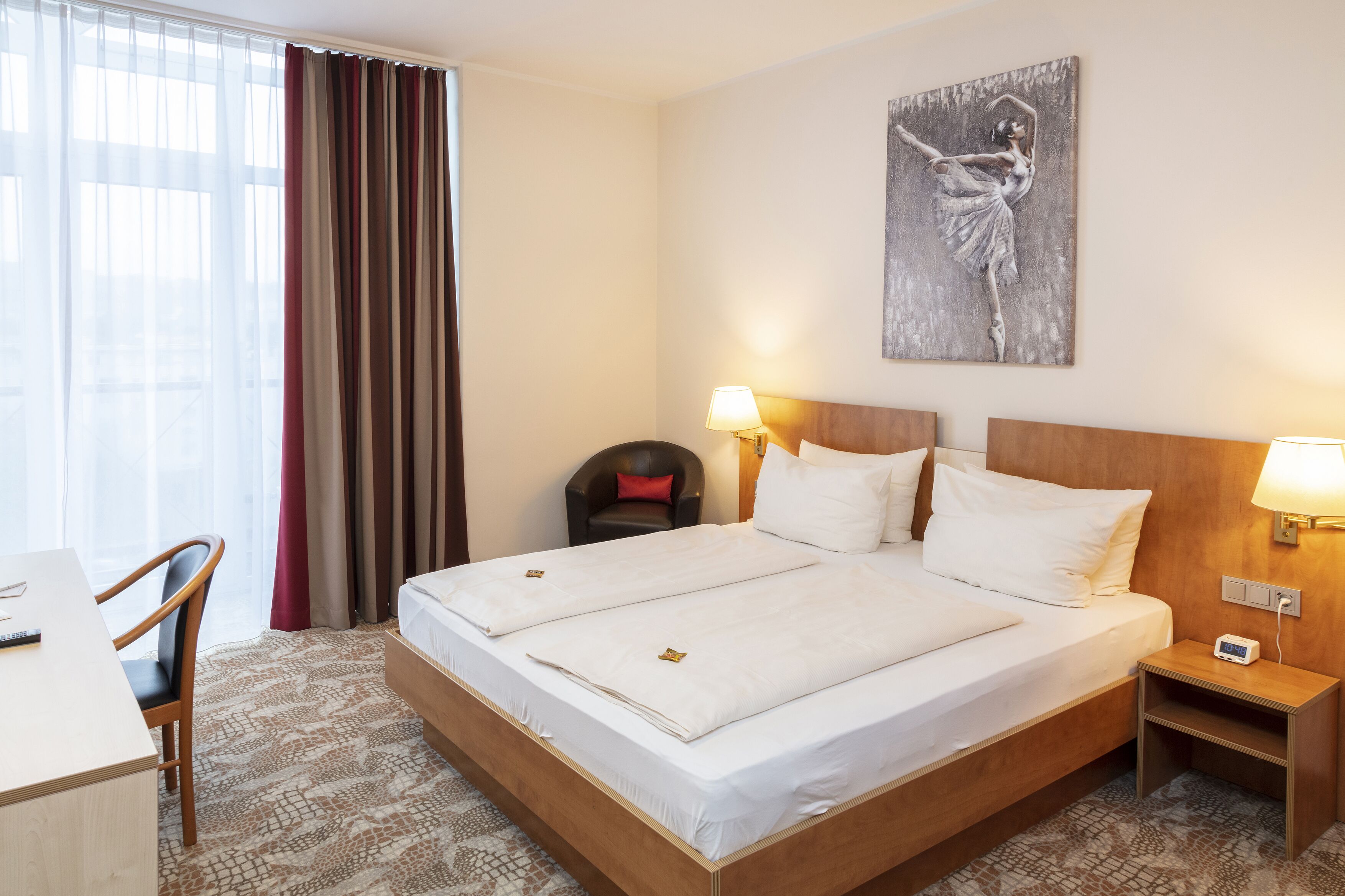 Ein Hotelzimmer mit einem großen Bett, zwei Nachttischen, einem Schreibtisch, einem Stuhl und einem Bild an der Wand.