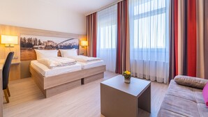 A modern hotel room with a bed, a desk, a chair, a sofa, and a view of a cityscape through large windows with sheer curtains. - Best Western Hotel Dreilaenderbruecke (Weil am Rhein)