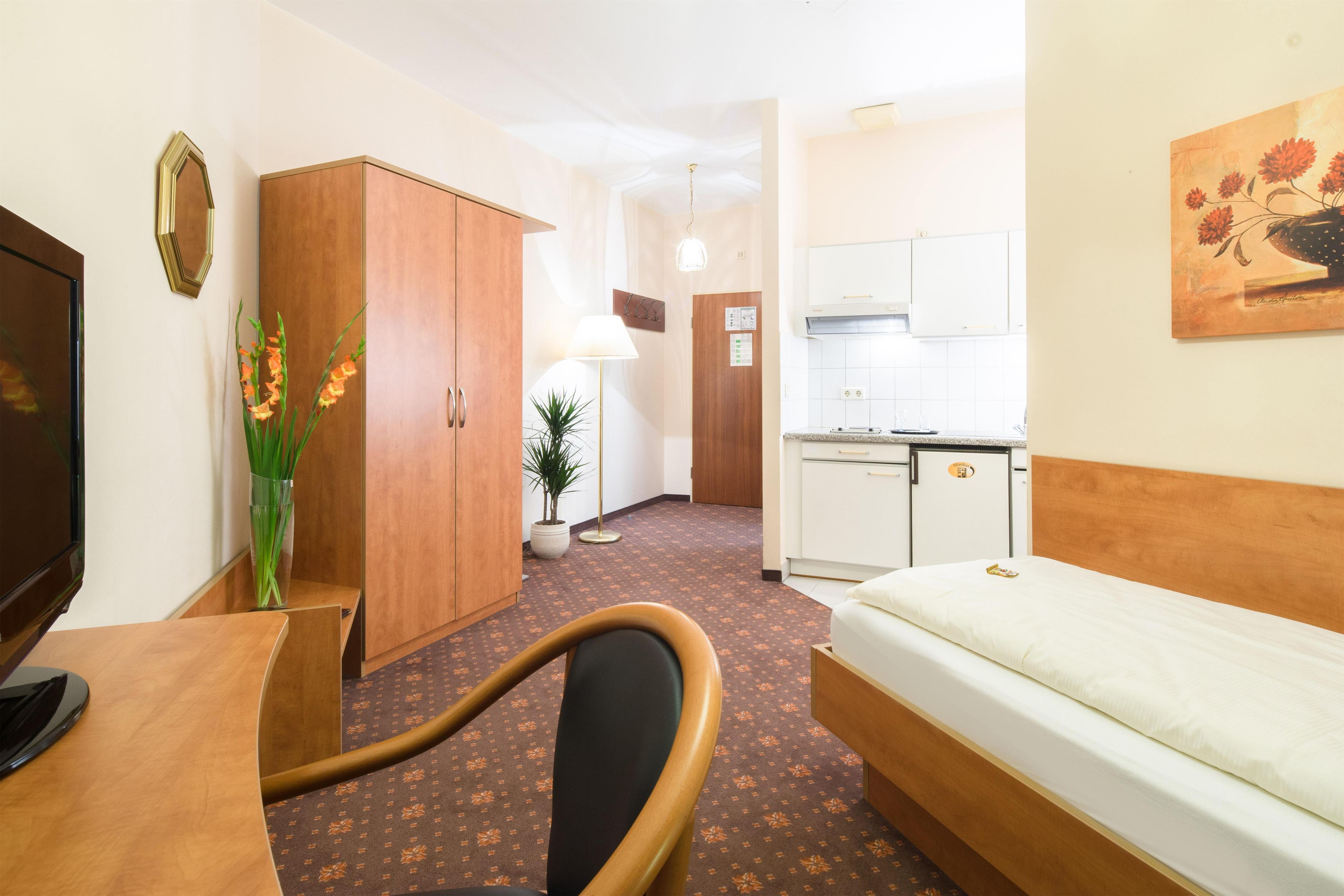 A hotel room with a bed, a desk, a chair, a wardrobe, a small kitchen area, and a potted plant.