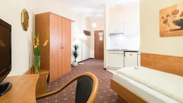 A hotel room with a bed, a desk, a chair, a wardrobe, a small kitchen area, and a potted plant.