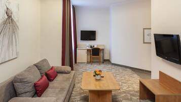 Una sala de estar amoblada con un sofá, una mesa de centro con un florero, un escritorio con una silla, un televisor de pantalla plana y una alfombra estampada.