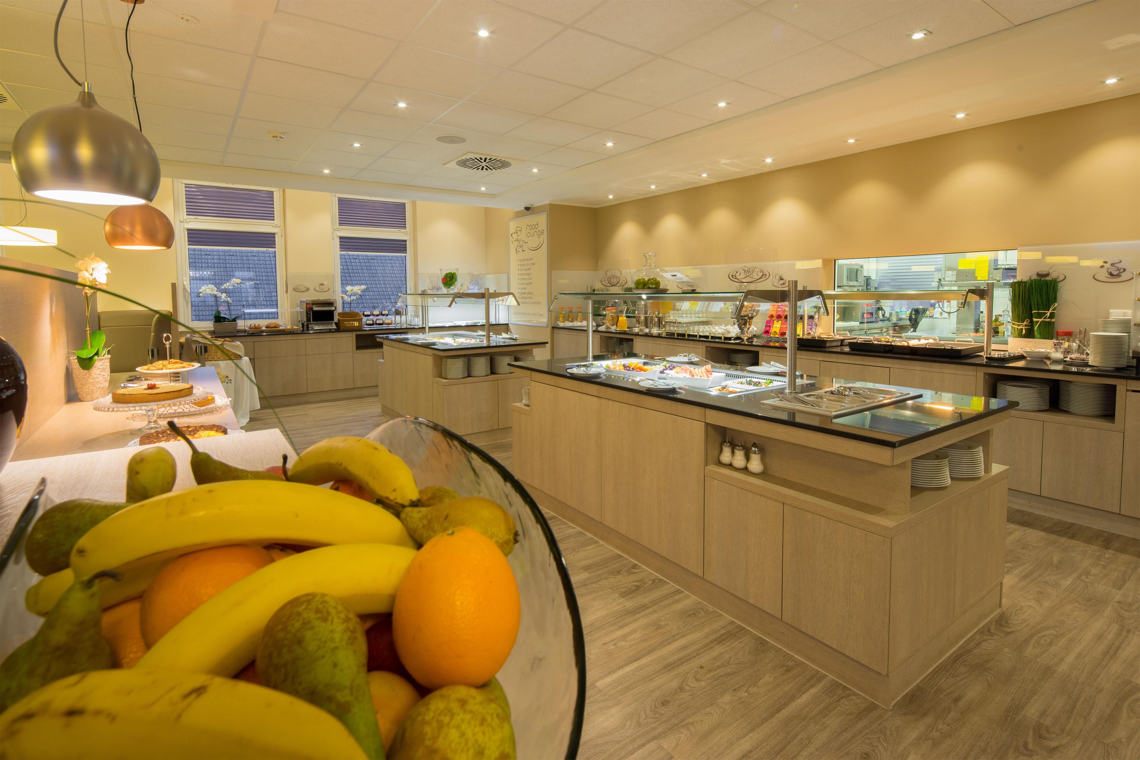A buffet area with a variety of food options, a fruit bowl in the foreground, and a view of the city through large windows.