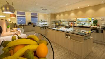 A buffet area with a variety of food options, a fruit bowl in the foreground, and a view of the city through large windows.