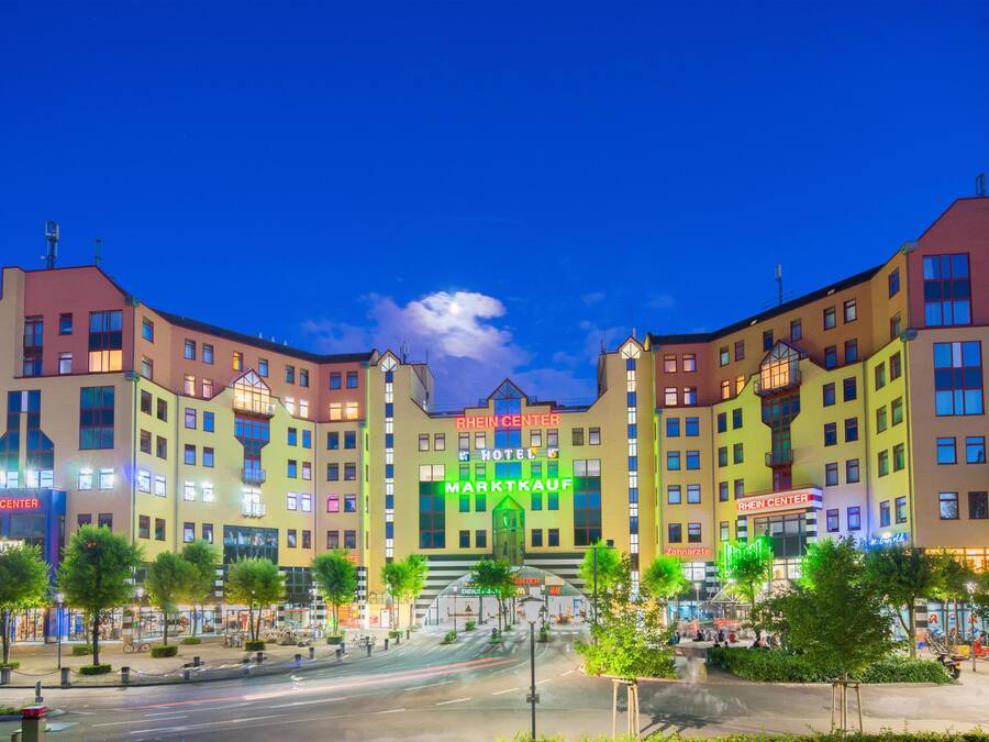 A multi-story hotel with a central entrance, surrounded by a landscaped area with trees and a fountain.