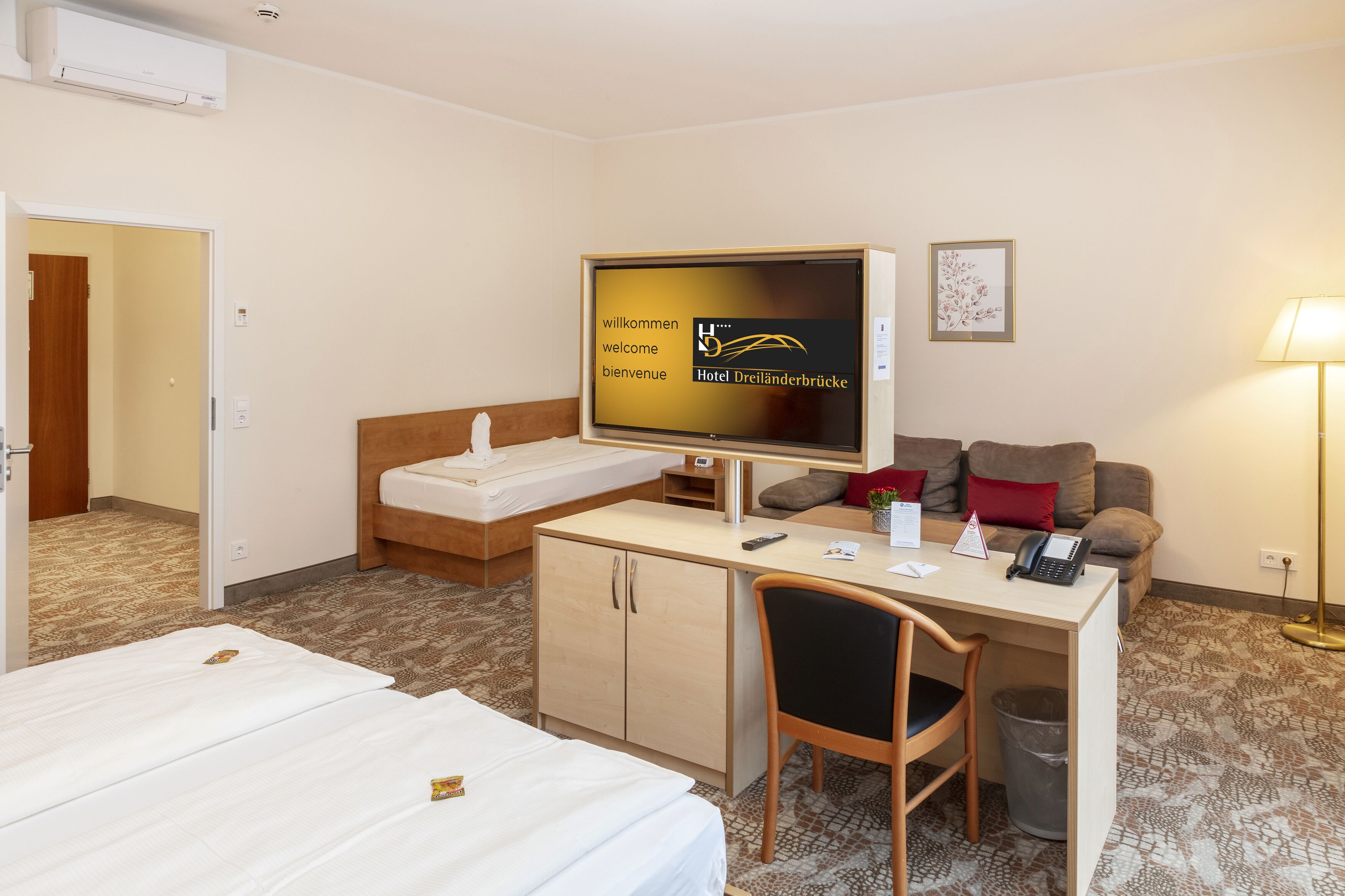 Une chambre d’hôtel équipée d’un lit, d’un bureau avec un ordinateur, d’une chaise, d’un canapé et d’un téléviseur affichant un message de bienvenue.