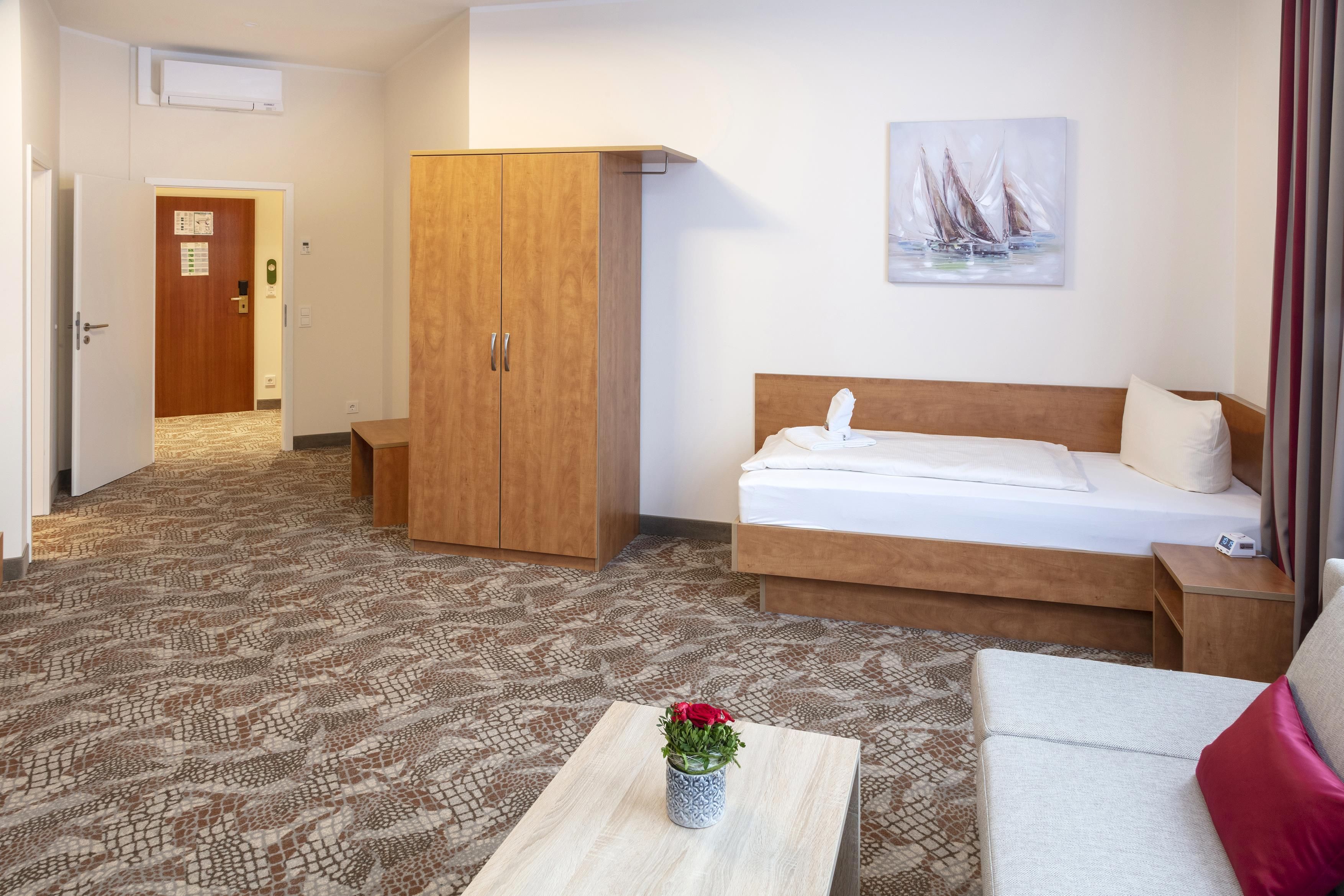 A hotel room with a bed, a wooden wardrobe, a small table with a vase of flowers, and a painting on the wall.