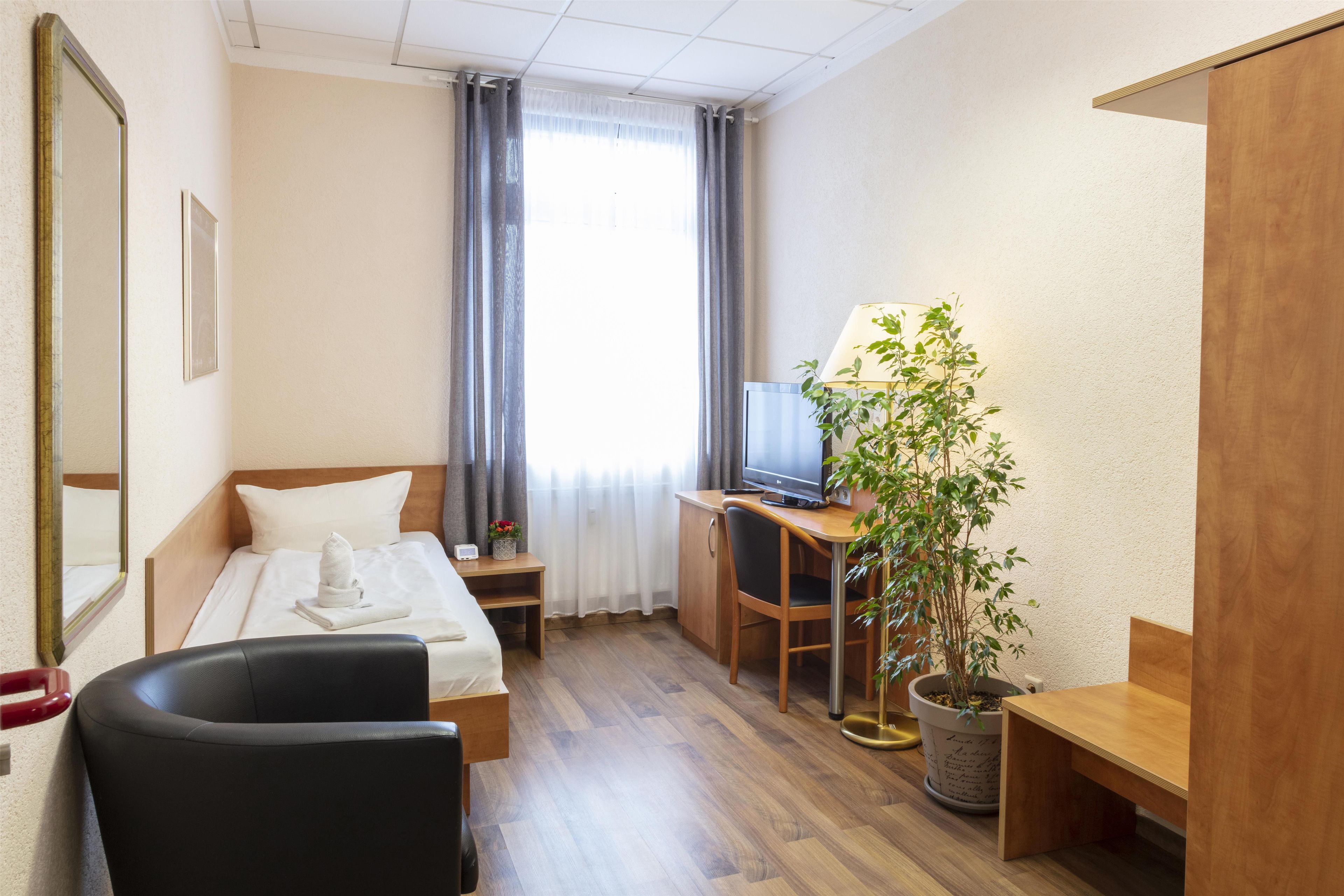 Une chambre d’hôtel avec un lit, un bureau avec un ordinateur, une chaise, une plante en pot et une fenêtre avec des rideaux.