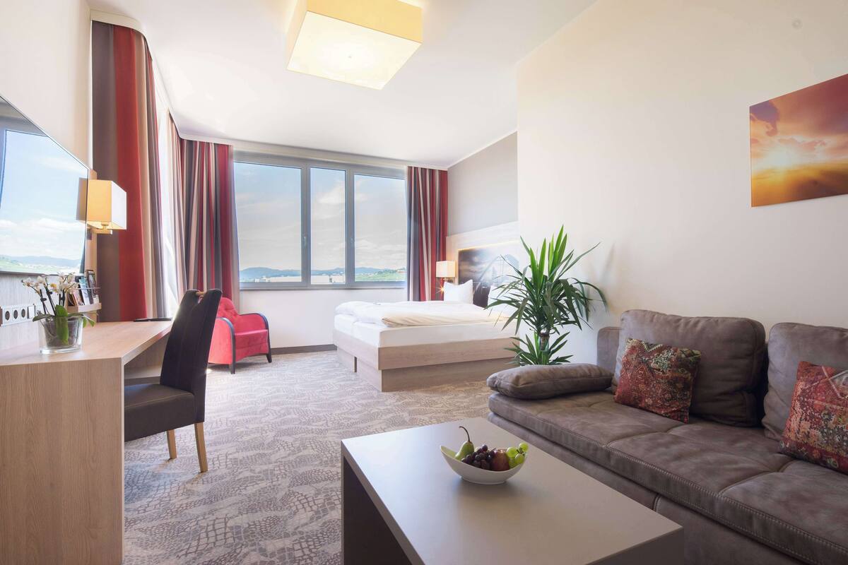 Una habitación de hotel moderna con cama, sofá, escritorio y un amplio ventanal con vistas a la ciudad.