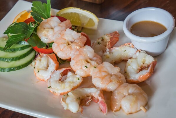A plate of shrimp with lemon slices, cucumber, and red bell pepper, accompanied by a small bowl of sauce.