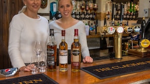 Two women standing behind a bar with various liquor bottles and glasses. - The Clachan Inn (Drymen)