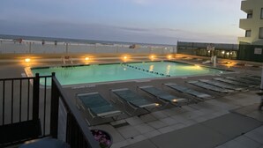 Une piscine sur le toit offrant une vue sur la plage et l’océan, entourée de chaises longues et d’un immeuble doté de balcons.