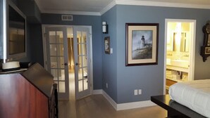 Une chambre à coucher avec un lit, une télévision, une porte donnant sur une salle de bain et un tableau accroché au mur.