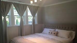 A bedroom with a large bed, a tufted headboard, and a view of the outdoors through the windows. - Lake Michigan Beach Cottages (Michigan City)