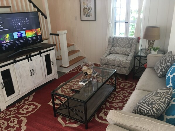 A living room with a TV, a patterned armchair, a glass coffee table, and a red patterned rug. - Lake Michigan Beach Cottages (Michigan City)