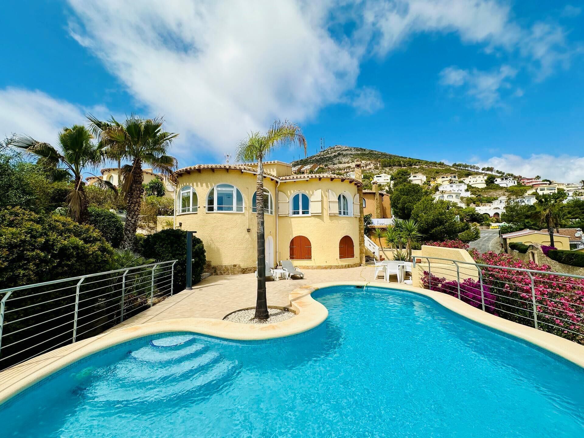 A pool area with a curved edge, a palm tree, and a building with arched windows and a tiled roof.