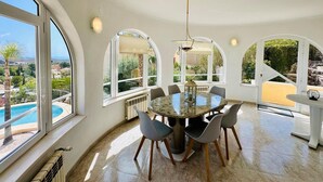 Un vaste espace dédié à la salle à manger, avec une table ronde et des chaises, de grandes fenêtres en arc de cercle et une vue sur une piscine.