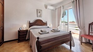 Ein Schlafzimmer mit einem hölzernen Bett, zwei Nachttischen, einem Schranken und einem Fenster mit Vorhängen, mit Blick ins Freie.