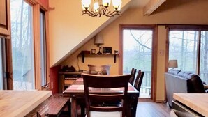 A dining area with a wooden table and chairs, a chandelier, and a view through large windows. - On the Betsie River, fish off dock, close to Crystal Mountain, trails, lakes! (Thompsonville)