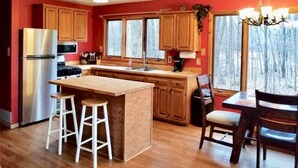 A kitchen with wooden cabinets, a stainless steel refrigerator, a wooden island with stools, and a window with a view of trees. - On the Betsie River, fish off dock, close to Crystal Mountain, trails, lakes! (Thompsonville)