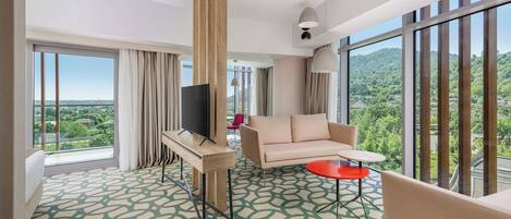 Una habitación de hotel moderna con un ventanal grande, un sofá, un televisor sobre un mueble bajo y una mesa roja.