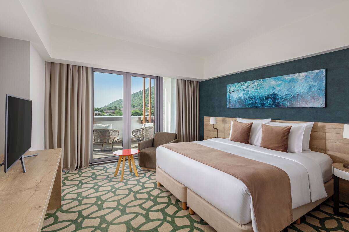 Ein Hotelzimmer mit einem großen Bett, einem Fernseher, einem Balkon mit Stühlen und einem Gemälde an der Wand.