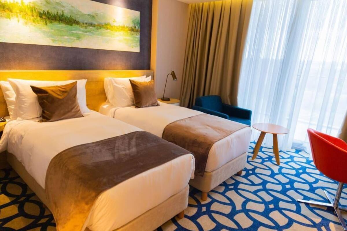 Ein Hotelzimmer mit zwei Betten, einem blauen Teppich mit Muster, einem roten Sessel und einem Bild an der Wand.