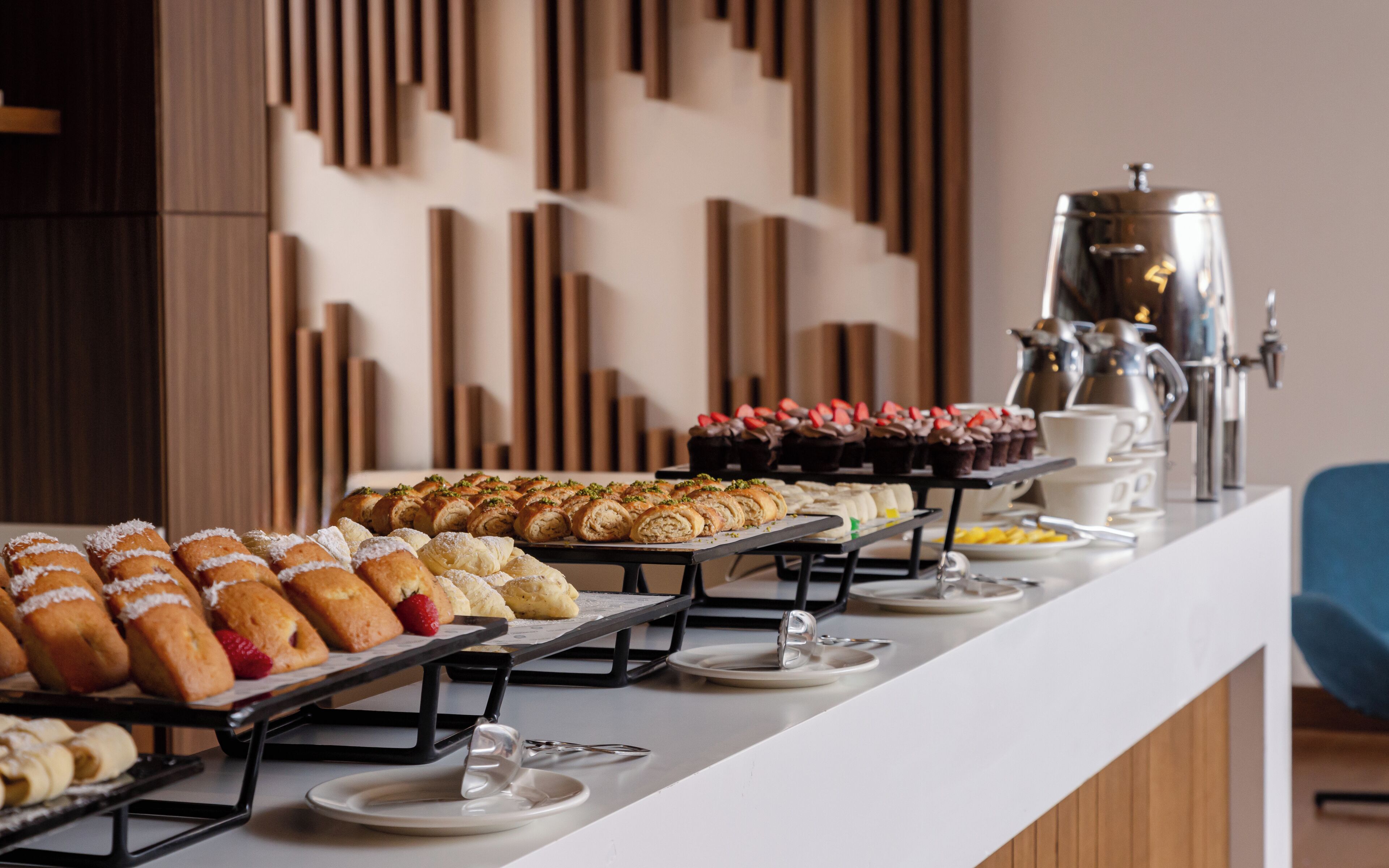 A buffet with various pastries and desserts, a coffee station, and a modern interior with vertical wooden panels.