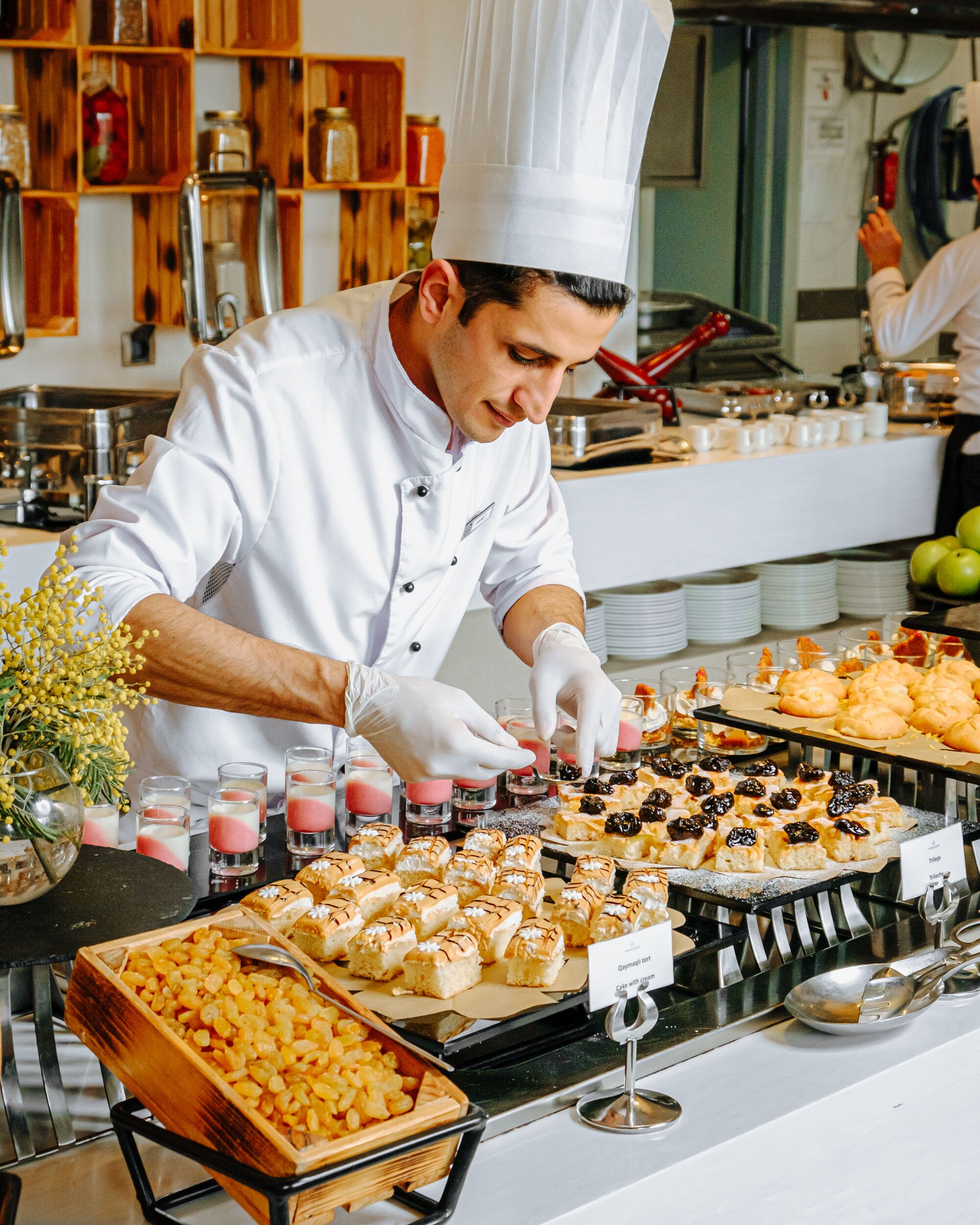 A chef in a white uniform preparing food in a kitchen with wooden shelves and various food items displayed on the counter.