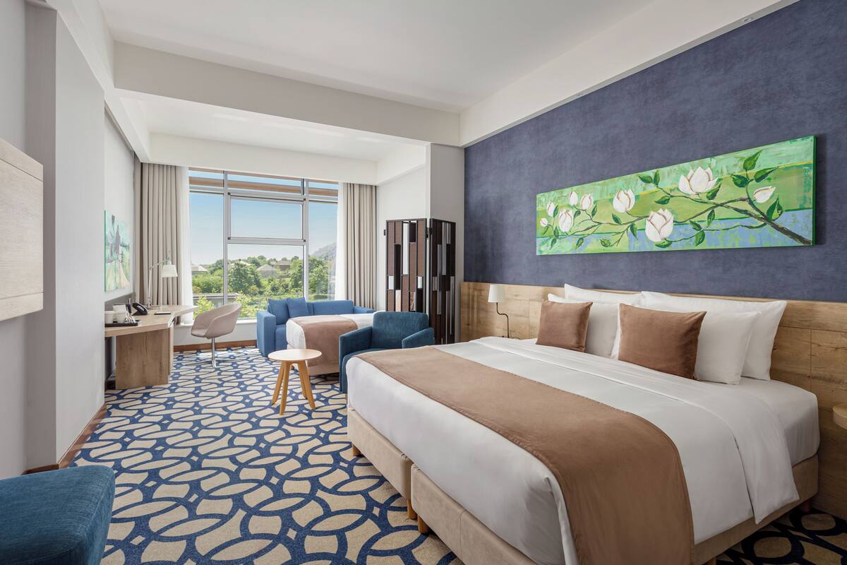 Ein Hotelzimmer mit einem großen Bett, einem Schreibtisch, einem Sessel und Blick ins Freie.
