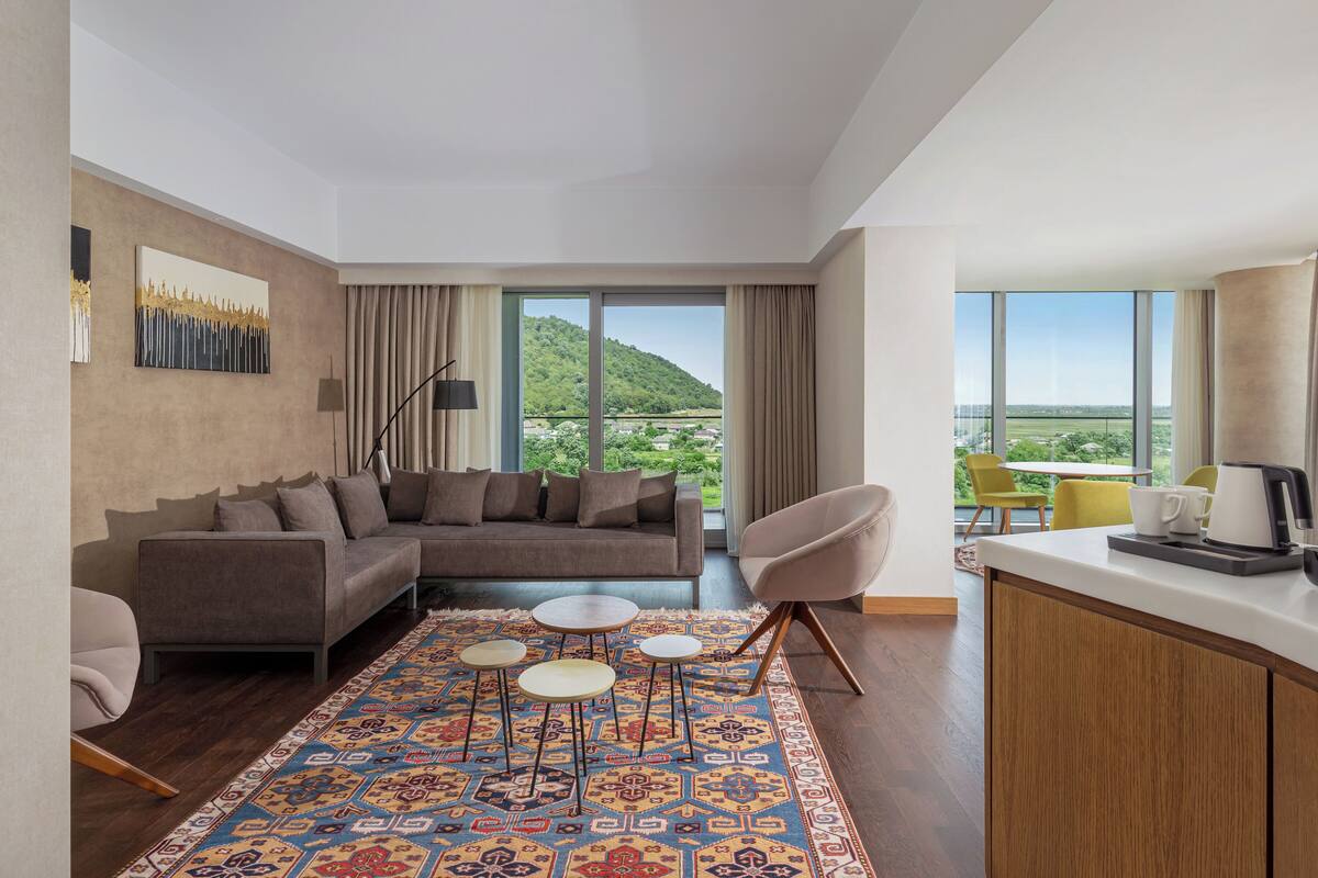 Ein modernes Hotelzimmer mit großem Fenster, einer Couch, einem Couchtisch, einem Sessel und einer Küchenzeile mit Kaffeemaschine.