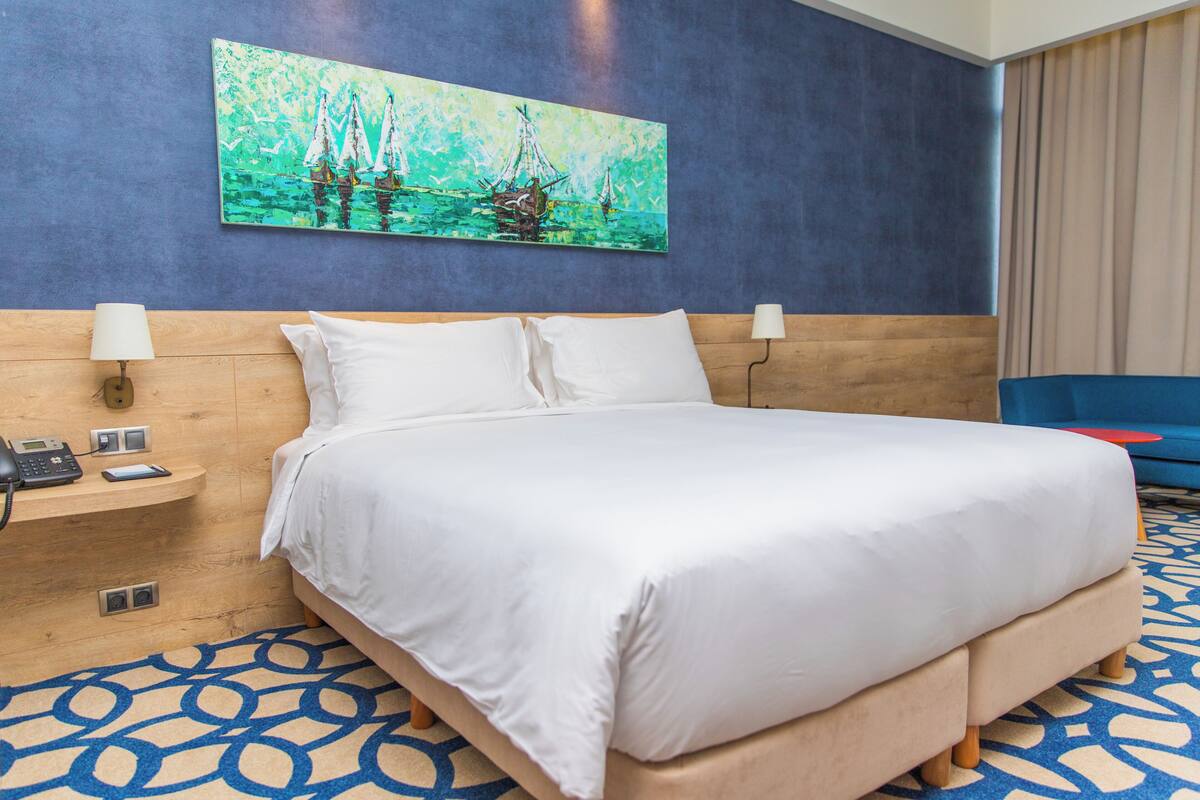 Ein Hotelzimmer mit einem großen Bett, einem hölzernen Kopfteil, einem Segelbootgemälde, einem blau gemusterten Teppich und einem blauen Sessel.