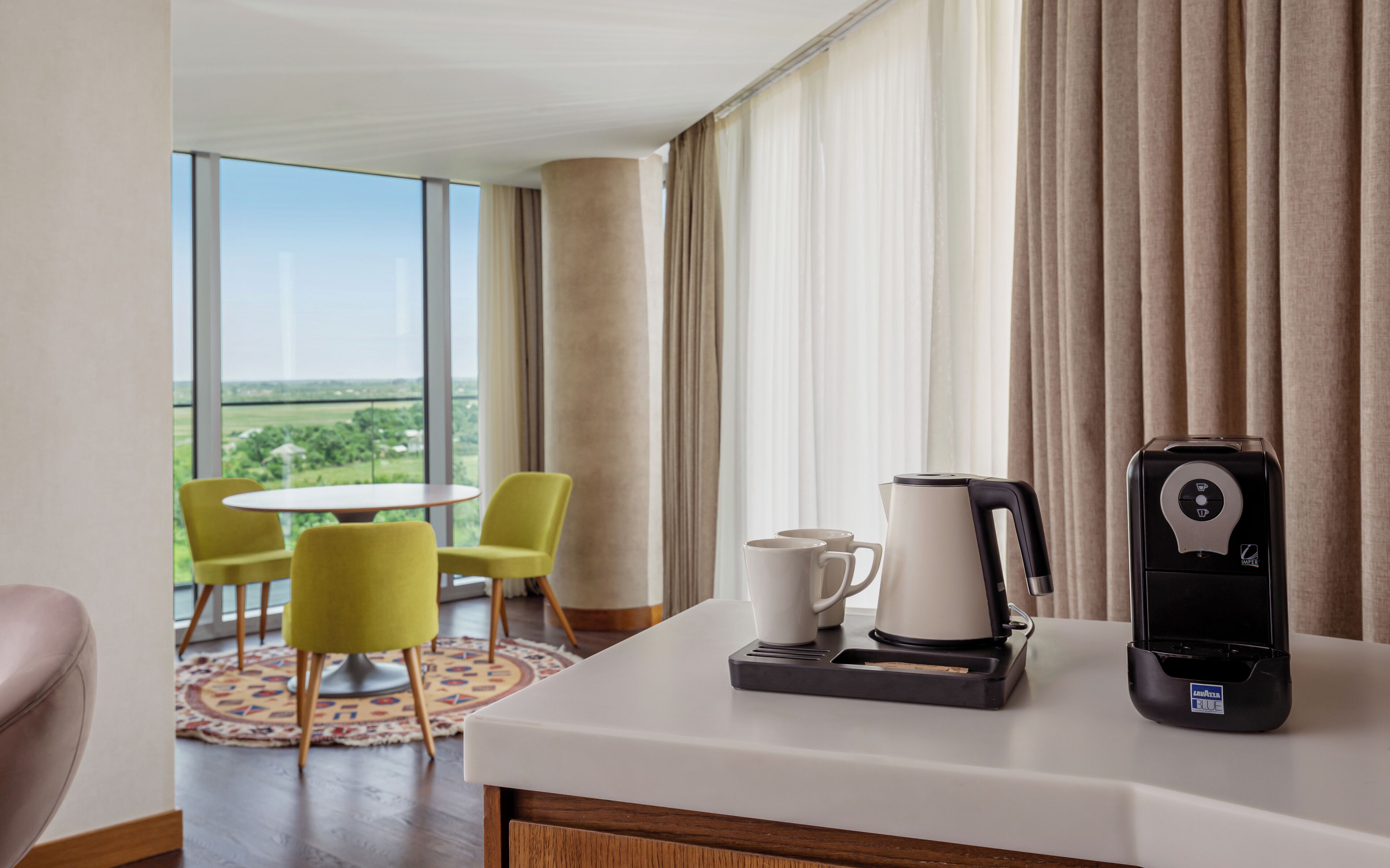 A hotel room with a large window, a coffee maker, a kettle, and two mugs on a table. There are two yellow chairs and a round table in the room.