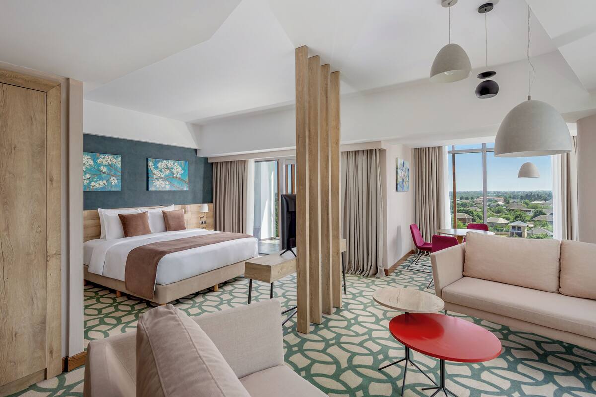 Ein modernes Hotelzimmer mit einem großen Bett, einer Sitzecke, einem roten Tisch und Blick auf die Stadt.