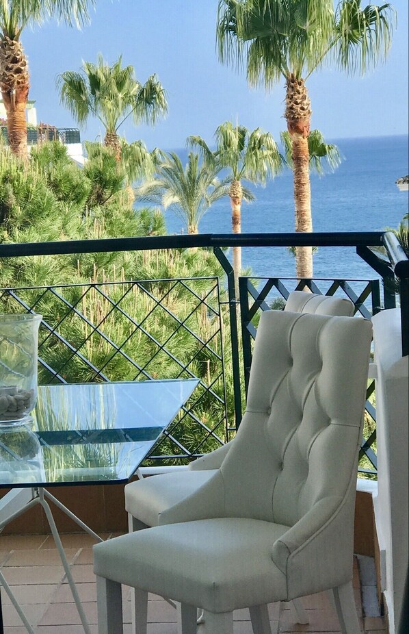 A balcony with a white cushioned chair and a glass table, overlooking a palm tree-lined view of the sea.