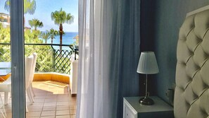 A balcony with a view of the sea and palm trees, a white chair, a table, and a lamp.