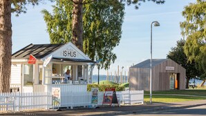 A small roadside kiosk with a white fence, a sign reading "ISHUS," and a view of the water in the background. - Apartments Bandholm (Bandholm)
