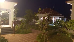 Vue nocturne d’un quartier résidentiel, avec un chemin pavé menant à une maison dont on aperçoit le porche et un petit jardin agrémenté de diverses plantes et d’un banc.