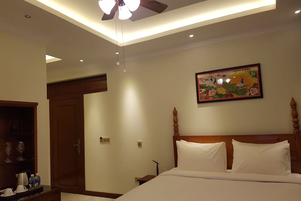 Ein Hotelzimmer mit Bett, Deckenventilator, Einbauleuchten, einem hölzernen Kopfteil, einem Nachttisch und einer Tür zu einem weiteren Raum.