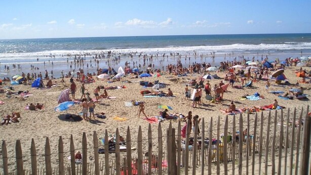 Una playa concurrida, con gente tomando el sol, nadando y paseando por la orilla.