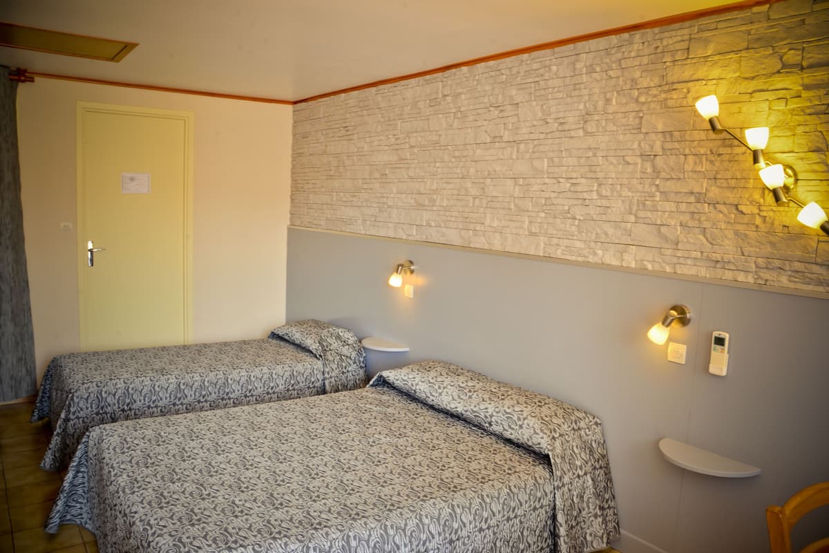 Une chambre avec deux lits, un mur en pierre, un luminaire et une petite étagère.