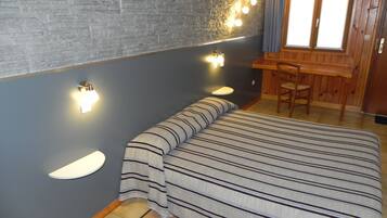 Une pièce comprenant un lit, une petite table et une chaise. Des lampes sont fixées au mur et il y a une fenêtre avec des rideaux.